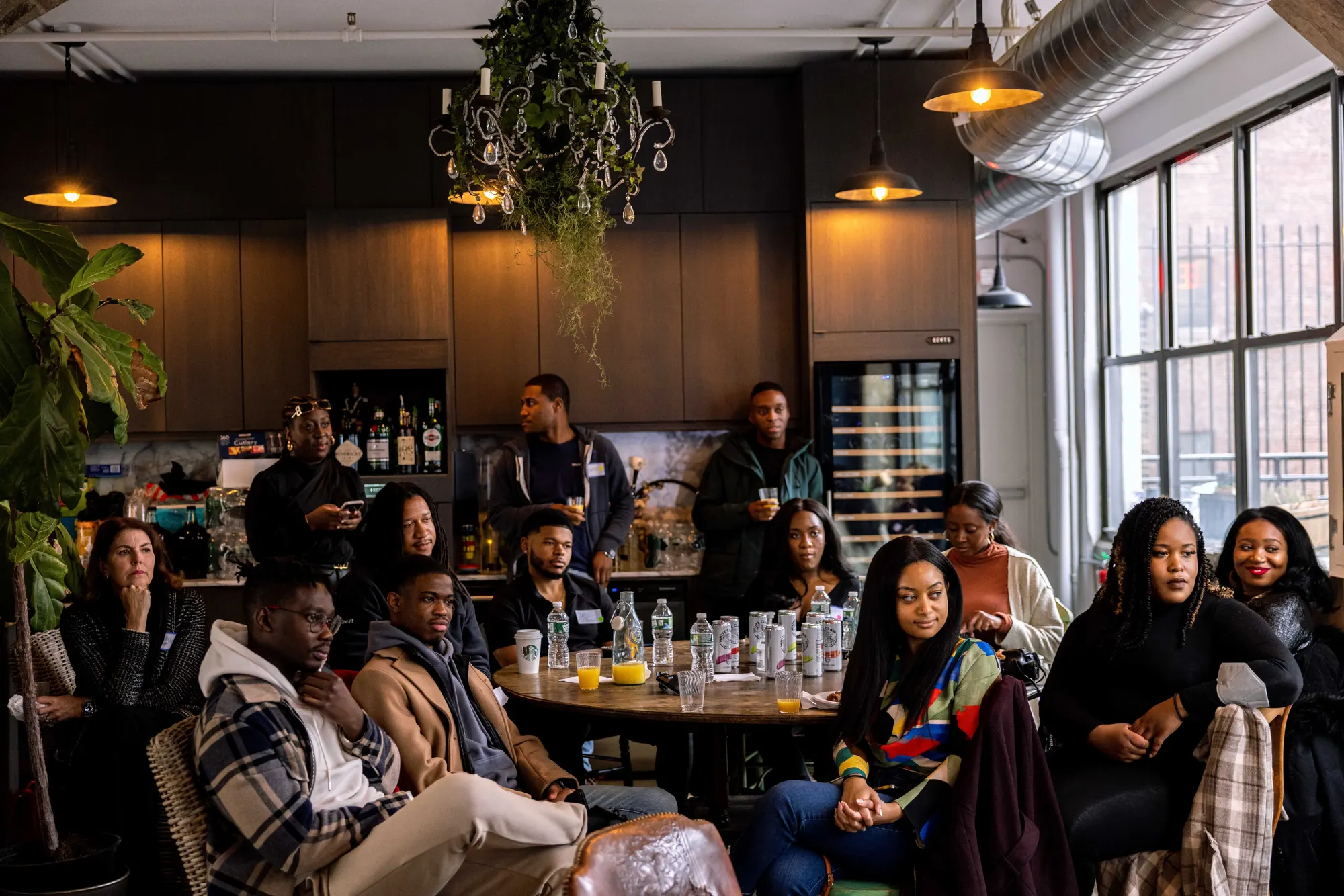 A crowded brunch in a loft, guests gathered under hanging plants and warm lamps
